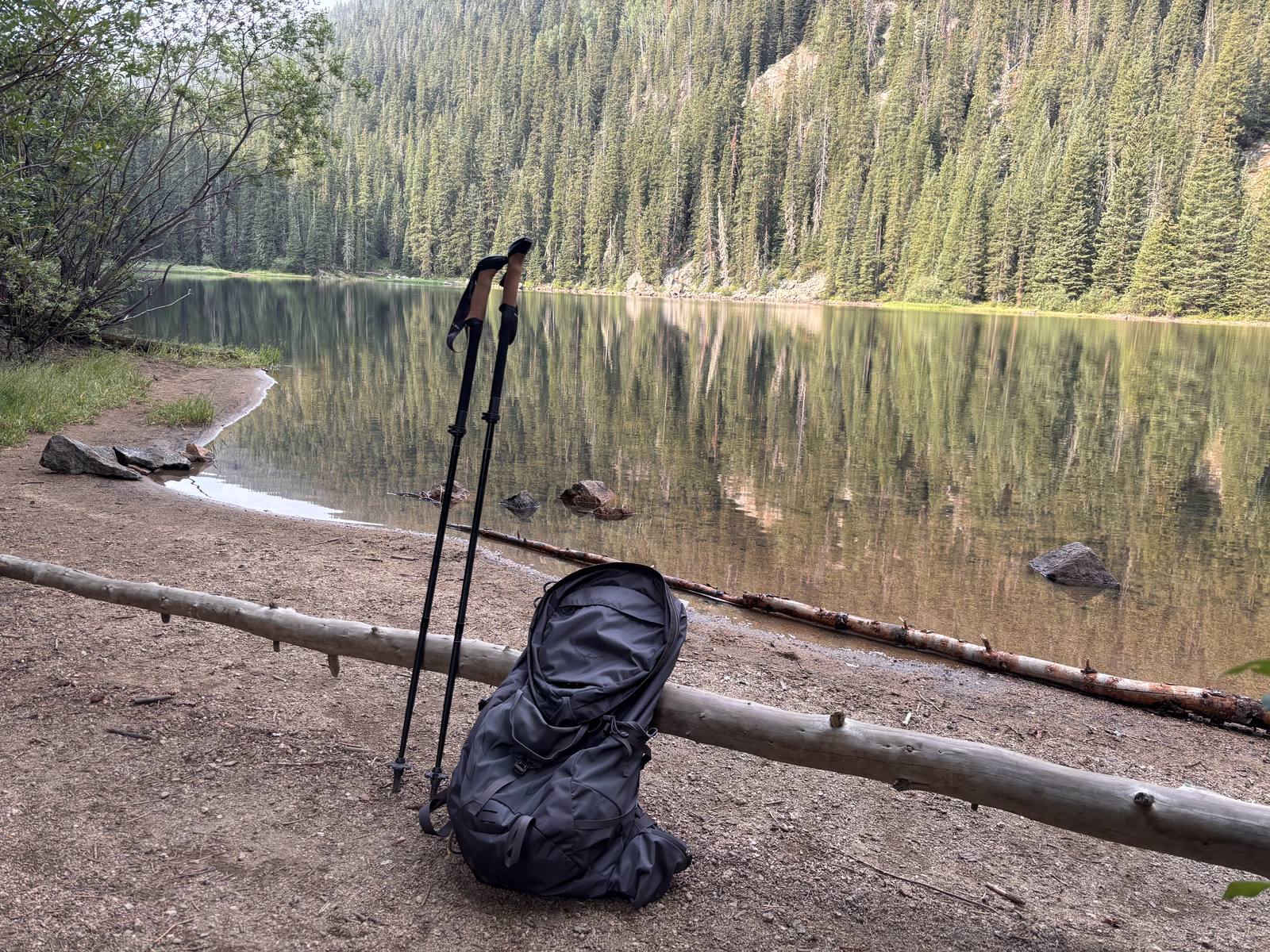 Hiking pack and trekking poles resting against a log on the shoreline of Beaver Lake in Beaver Creek, with forested slopes reflected in still water